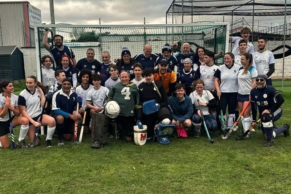 Bexleyheath & Belvedere Hockey Club players pose in the goal for the team photo. The youngsters are in white. The veterans are in blue. This pitch is on grass. 
